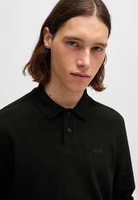 Camisa polo de manga longa preta, feita de tecido texturizado. Apresenta um colarinho dobrado, botões duplos na frente e um logótipo em relevo no peito.