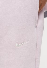 Κοντινή λήψη ροζ παντελονιού φόρμας Nike με λευκό λογότυπο Nike swoosh κεντημένο κοντά στην πλαϊνή τσέπη όπου τοποθετείται το χέρι.