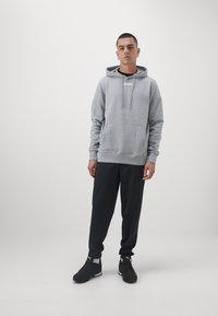 Felpa grigia con tasca frontale e cappuccio regolabile, abbinata a pantaloni sportivi neri e sneakers nere. Design semplice e casual.