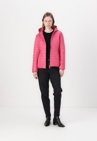 Pink quilted jacket with a hood, front zipper, and textured lines, worn over a black top and paired with black pants and ankle boots.