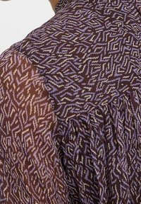 Indumento a maniche lunghe trasparente con motivo a zigzag astratto in viola e bianco su sfondo marrone scuro.