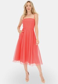 Koronkowa sukienka midi w kolorze koralowym z cienkimi ramiączkami, dopasowanym korpusem i falującą, przezroczystą spódnicą. Posiada boczne kieszenie i gładką teksturę.