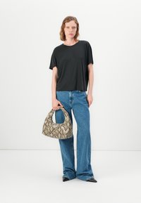 Kvinne i svart t-skjorte og blå jeans som holder en beige håndveske med slangeskinnmønster mot hvit bakgrunn.