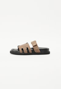 Suede glidesandal i brun og svart. Har to brede utskjæringer på overdelen og et justerbart belte med en gullspenne. Flat såle.