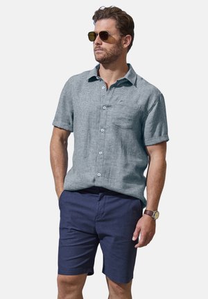 Man wearing textured short-sleeve gray button-up shirt, navy shorts, brown watch, and sunglasses, standing with hands in pockets on white background.