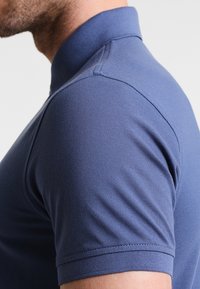 Námořnická polo tričko s detailním záběrem na límec a rameno, s texturovanou tkaninou a krátkými rukávy s pečlivým prošitím.