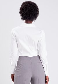 Chemise blanche à manches longues avec col, texture lisse et poignets à boutons, associée à un pantalon gris ajusté, vue de dos.