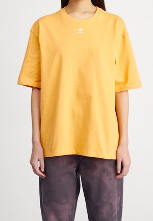 Żółta bawełniana koszulka z okrągłym dekoltem, krótkimi rękawami i białym logo na piersi, zestawiona z ciemnymi spodniami w wzory.