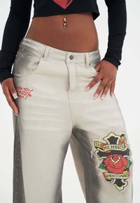 Pantalones de mezclilla de color claro con un acabado desgastado. Presenta texto bordado en rojo y un gran parche con un diseño de rosa y texto en el muslo.
