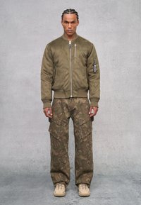 Chaqueta bomber de ante verde con cierre de cremallera, combinada con pantalones cargo de camuflaje que cuentan con grandes bolsillos. Zapatillas beige completan el conjunto.