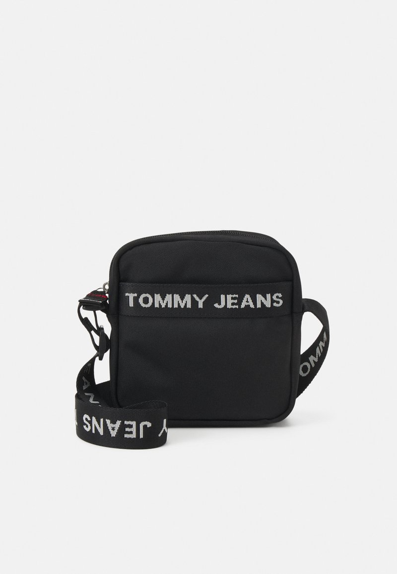 Bolso de hombro rectangular negro fabricado en tela, con cierre de cremallera y el logo "TOMMY JEANS" en blanco en la parte frontal y la correa.