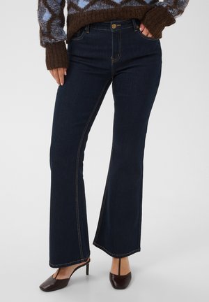 Personne portant un jean bootcut bleu foncé, des chaussures à talons marron et un pull marron et bleu à motifs, une main dans la poche.