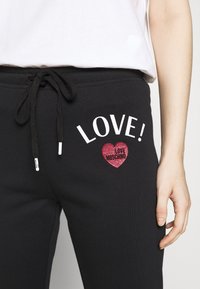 Black sweatpants featuring a drawstring waist, white "LOVE!" text, and a glittery red heart with "LOVE MOSCHINO" text. Smooth cotton fabric.