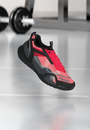 Lekki różowo-czarny but sportowy Under Armour zawieszony nad podłogą siłowni, z rozmytymi hantlami w tle.