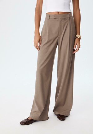Femme portant un pantalon beige à rayures à taille haute et jambes larges, un haut blanc sans manches, des chaussures plates marron et un bracelet en or au poignet gauche.