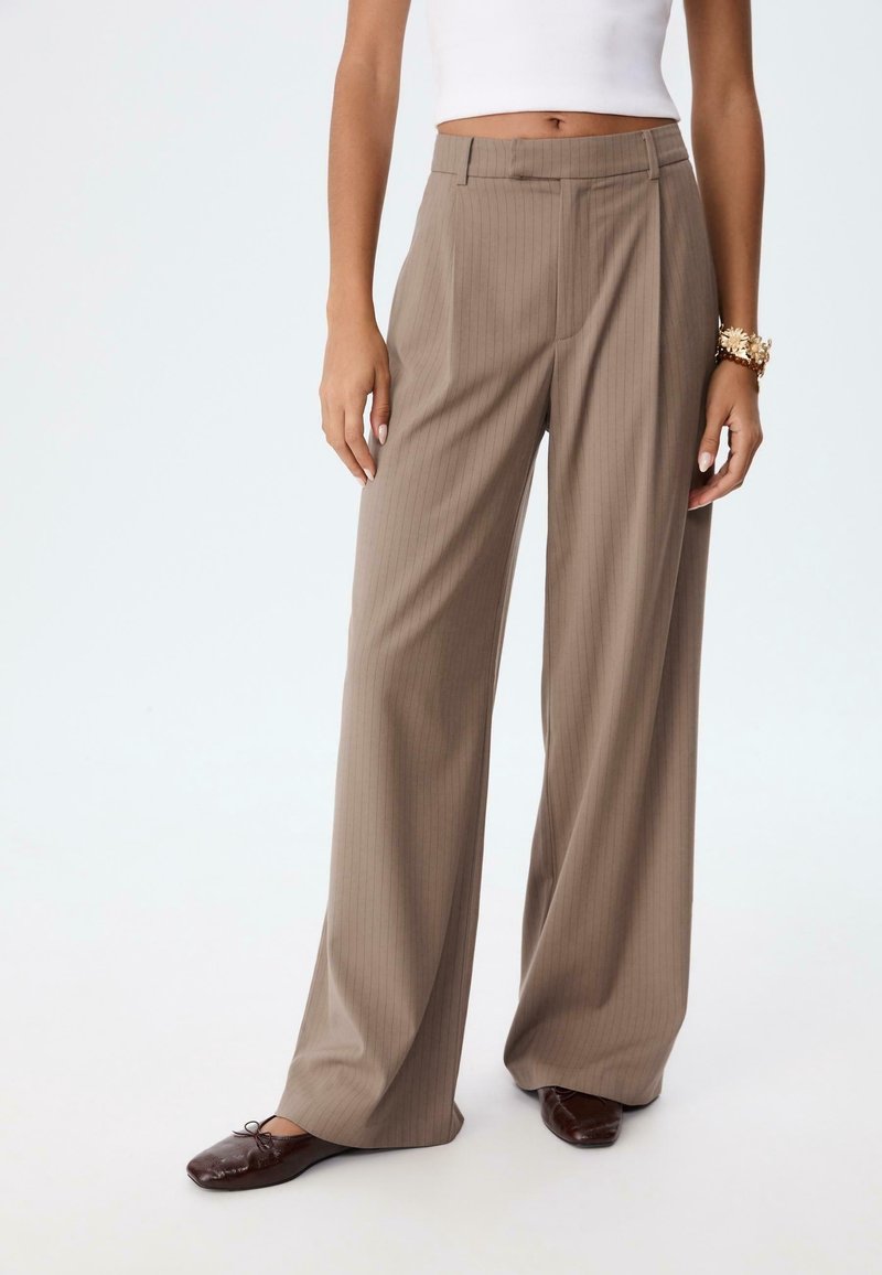 Femme portant un pantalon beige à rayures à taille haute et jambes larges, un haut blanc sans manches, des chaussures plates marron et un bracelet en or au poignet gauche.