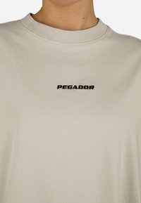 Gaiši pelēkas kokvilnas T-krekls ar apvalku un melnu izšūtu "PEGADOR" logotipu uz krūtīm. Gluda tekstūra un brīva piegriezuma. 