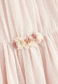 Jasnoróżowa sukienka z fakturowanej, kropkowanej tkaniny. Posiada marszczoną talię ozdobioną warstwowymi kwiatami z tkaniny w odcieniach różu i kremu.