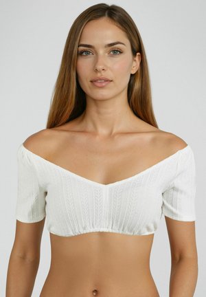 Jeune femme aux longs cheveux bruns portant un haut court en maille blanche à épaules dénudées, sur un fond gris uni.