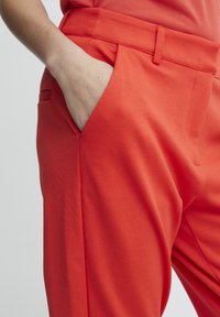 Red cotton trousers with a smooth texture, featuring side pockets and a tailored cut, showcasing sharp pleats at the waist.