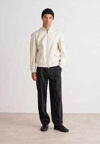 White bomber jacket made of smooth material with a front zip, paired with black trousers and black sneakers, featuring minimalistic design.