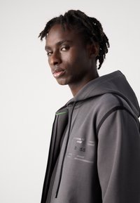 Felpa grigia con cappuccio realizzata in tessuto morbido, dotata di un logo nero, dettagli e accenti, e un tocco di verde sulla zip. Design moderno con vestibilità slim.