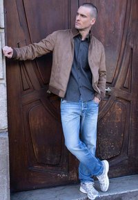 Chaqueta bomber de cuero marrón sobre una camisa de botones oscura, acompañada de unos vaqueros azul claro y zapatillas deportivas grises. De pie contra una puerta de madera.