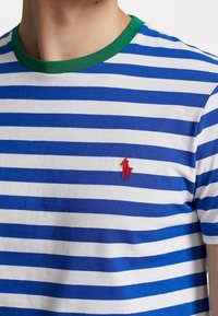 Modré a bílé pruhované tričko s zeleným límečkem, vyrobené z bavlny, s malým červeným vyšitým logem na levé straně hrudi.
