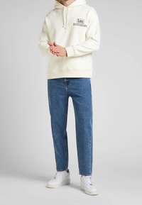 Sudadera crema con logo frontal, combinada con jeans de denim azul rectos y zapatillas blancas. Diseño simple con cordón y puños acanalados.