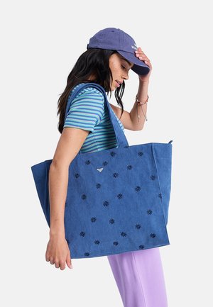 Kobieta ubrana w niebiesko-zieloną koszulę w paski, fioletowe spodnie i niebieską czapkę, trzymająca dużą niebieską torbę na ramię z czarnymi kwiatowymi wzorami.