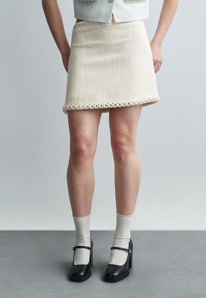 Femme portant une mini-jupe crème texturée avec bordure en perles, chaussettes beiges et talons Mary Jane noirs, debout sur un sol gris.