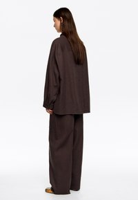 Camisa marrón de gran tamaño y pantalones anchos a juego, confeccionados en una tela texturizada. El conjunto presenta un corte relajado y detalles de botones.