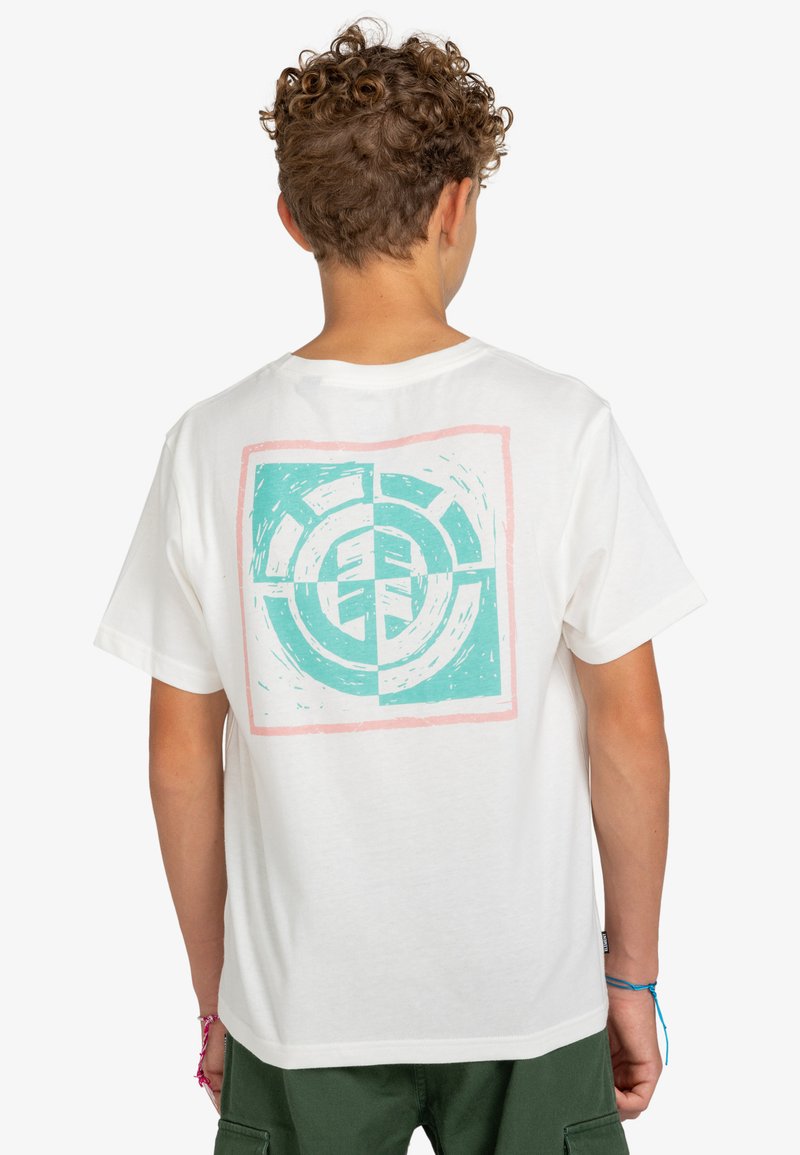 Jong persoon met krullend haar, gekleed in een wit T-shirt met een abstract cirkelontwerp in teal en roze op de achterkant en groene broek.
