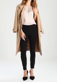 Beige trenchcoat over en lys bluse, parret med sorte tætsiddende bukser og sorte hæle. Frakken har et dobbeltradet design og hakkerlapper.