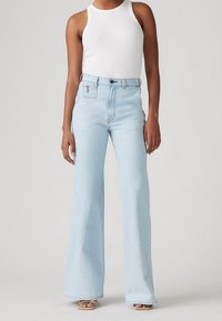 Person wearing high-waisted, light blue flare jeans, white sleeveless top, and beige open-toe sandals on a plain background.