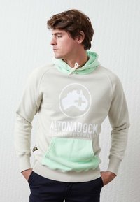 Sudadera beige clara con capucha y bolsillo en verde menta, con un logo y texto en blanco. Tejido suave, corte casual, mangas largas.