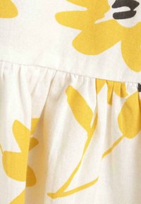 Tejido floral con grandes flores amarillas y tallos verdes sobre un fondo blanco, con un detalle de costura fruncida en la parte inferior.