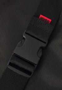 Boucle en plastique noir fixé sur une sangle tissée noire avec une petite étiquette rouge portant le label "HUGO" sur un fond de tissu texturé noir.