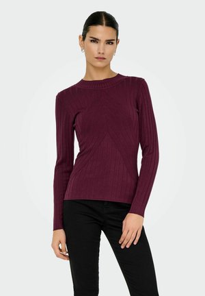Vrouw met steil, donker haar draagt een aansluitende, geribbelde, lange mouwen bordeauxrode trui en zwarte broek, staande tegen een effen witte achtergrond.