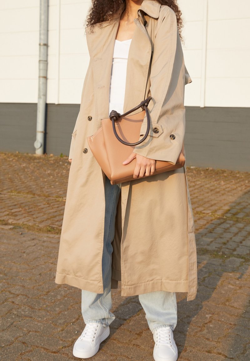 Femme portant un trench beige, un jean bleu clair, des baskets blanches, tenant un sac à main beige avec une poignée foncée, debout sur un trottoir pavé.