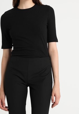 Femme portant un top noir côtelé ajusté à manches courtes et un pantalon noir taille haute sur un fond clair uni.