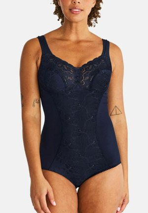Vrouw draagt marineblauwe kanten body met brede bandjes, staand met zichtbare armtattoeages tegen een witte achtergrond.