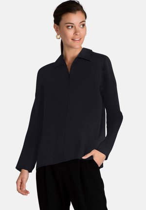 Blouse noire à manches longues avec un col et un détail de fermeture éclair à l'avant. Texture lisse et coupe décontractée, agrémentée de poches latérales.