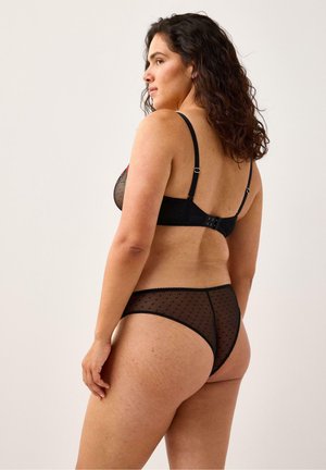 Musta alusvaatesett, jossa on läpinäkyvä kangas ja pilkkukuosi, säädettävät olkaimet ja takana hakaskiinnitys.