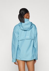 Donna con coda di cavallo scura, che indossa una giacca con cappuccio azzurro chiaro e pantaloni corti a righe, rivolta di spalle su uno sfondo bianco uniforme.