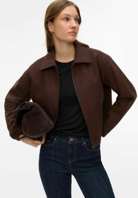 Jeune femme portant une veste marron zipp ée et un jean foncé, tenant un sac à main plié assorti marron, regardant sur le côté.