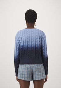 Kabelstickad tröja i graduerad blå, som övergår från ljus till mörk. Har en ribbad krage och nederkant, i kombination med rutiga denimshorts.