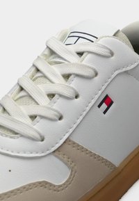 Λευκό αθλητικό παπούτσι με μπεζ λεπτομέρεια, επάνω μέρος από υφή δέρματος, επίπεδα λευκά κορδόνια, ετικέτα λογότυπου και ελαστική σόλα με ελαφρύ ανάγλυφο μοτίβο.