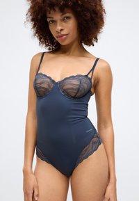 Sötétkék bodysuit csipkés kosarakkal, fodros szélekkel és vékony spagetti pántokkal. Sima anyagból készült, alakra simuló szabással. Calvin Klein brandinggel.