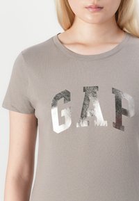 Young person wearing taupe t-shirt with large metallic silver "GAP" logo across chest, cropped mid-face and arm.